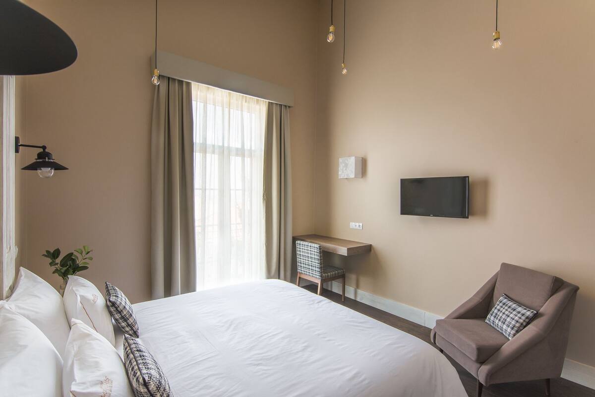 A hotel room with a large bed, a desk, a chair, a TV, and a window with curtains.