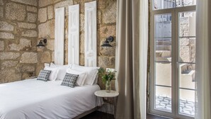 A bedroom with a stone wall, a bed with white bedding, two wall-mounted lamps, and a window with curtains.