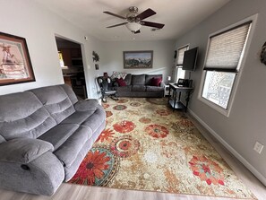Un salon avec un canapé gris, un tapis à motifs, un ventilateur de plafond, un téléviseur et une fenêtre avec des stores.