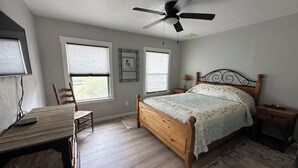 Un dormitorio con cama de madera, escritorio, silla, ventilador de techo y dos ventanas con persianas.