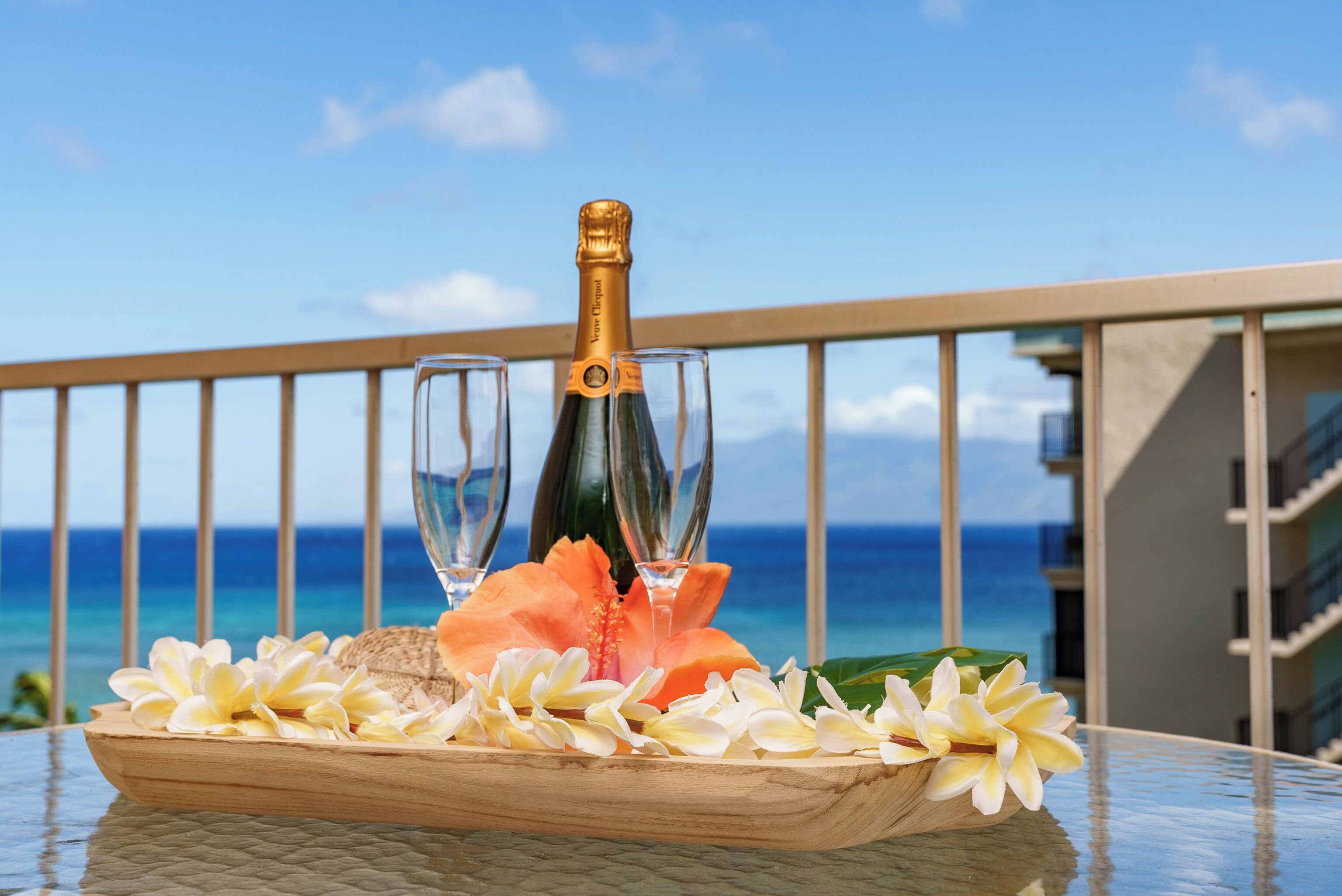 En flaska champagne, två glas och ett träbricka med blommor på en balkong med utsikt över havet.