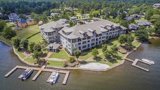 An aerial view of a large, multi-story building with a waterfront dock, surrounded by lush greenery and other residential structures.