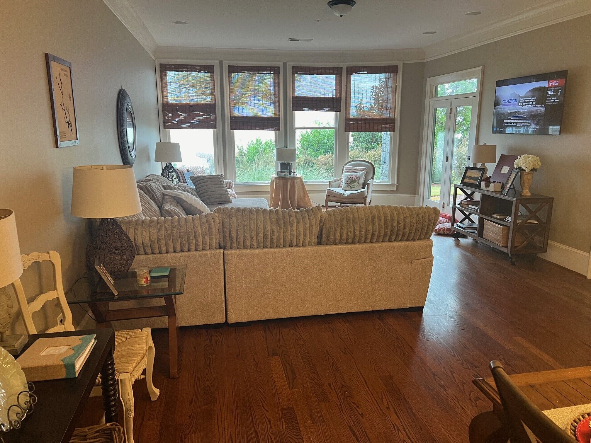 A living room with a beige sectional sofa, a small table with a lamp, a chair, and a TV mounted on the wall.