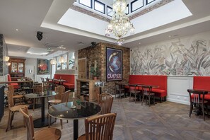Een restaurant met hoge plafonds, een kroonluchter en een muurschildering van een planeet. Tafels en stoelen staan er klaar voor dineren.