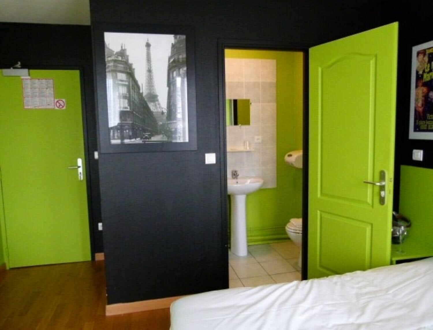 Una habitación con una cama, una puerta verde, una pared negra y un cuadro enmarcado de un paisaje urbano.