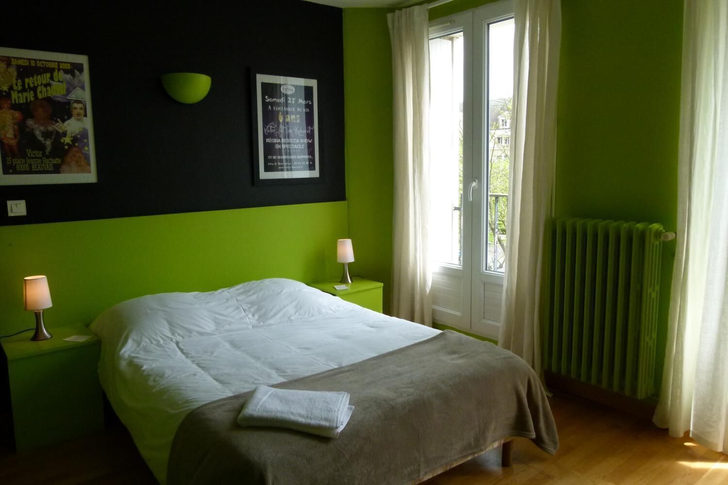 Una habitación con una cama, dos mesitas de noche con lámparas, un cartel en la pared y una ventana con cortinas.