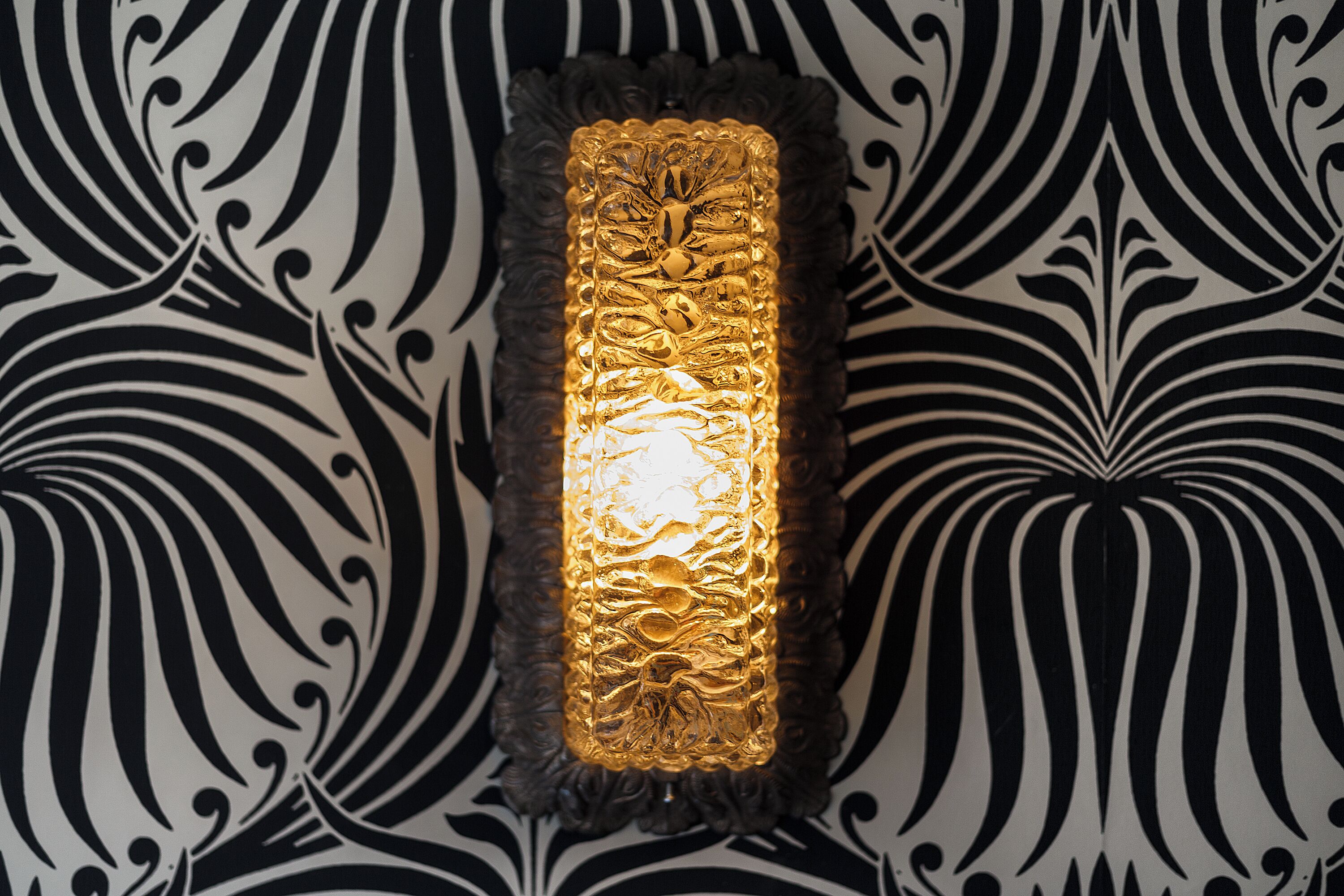 A wall-mounted light fixture with a textured, gold-colored design, set against a black and white patterned wallpaper.