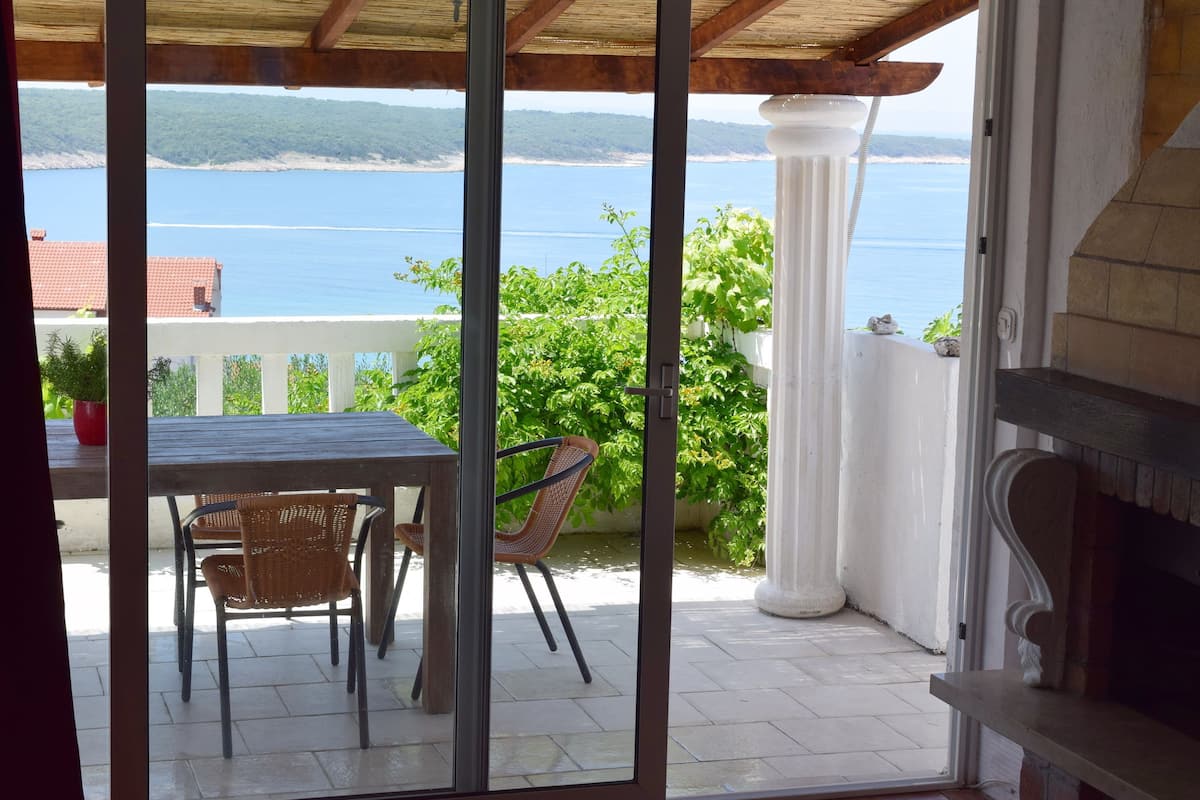 A covered patio with a table and chairs, a view of the sea, and a white column.
