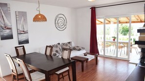 A dining area with a wooden table and chairs, a sofa with patterned cushions, and a large window with a view of the outdoors. - Villa Agata Rab - Apartments (Rab)