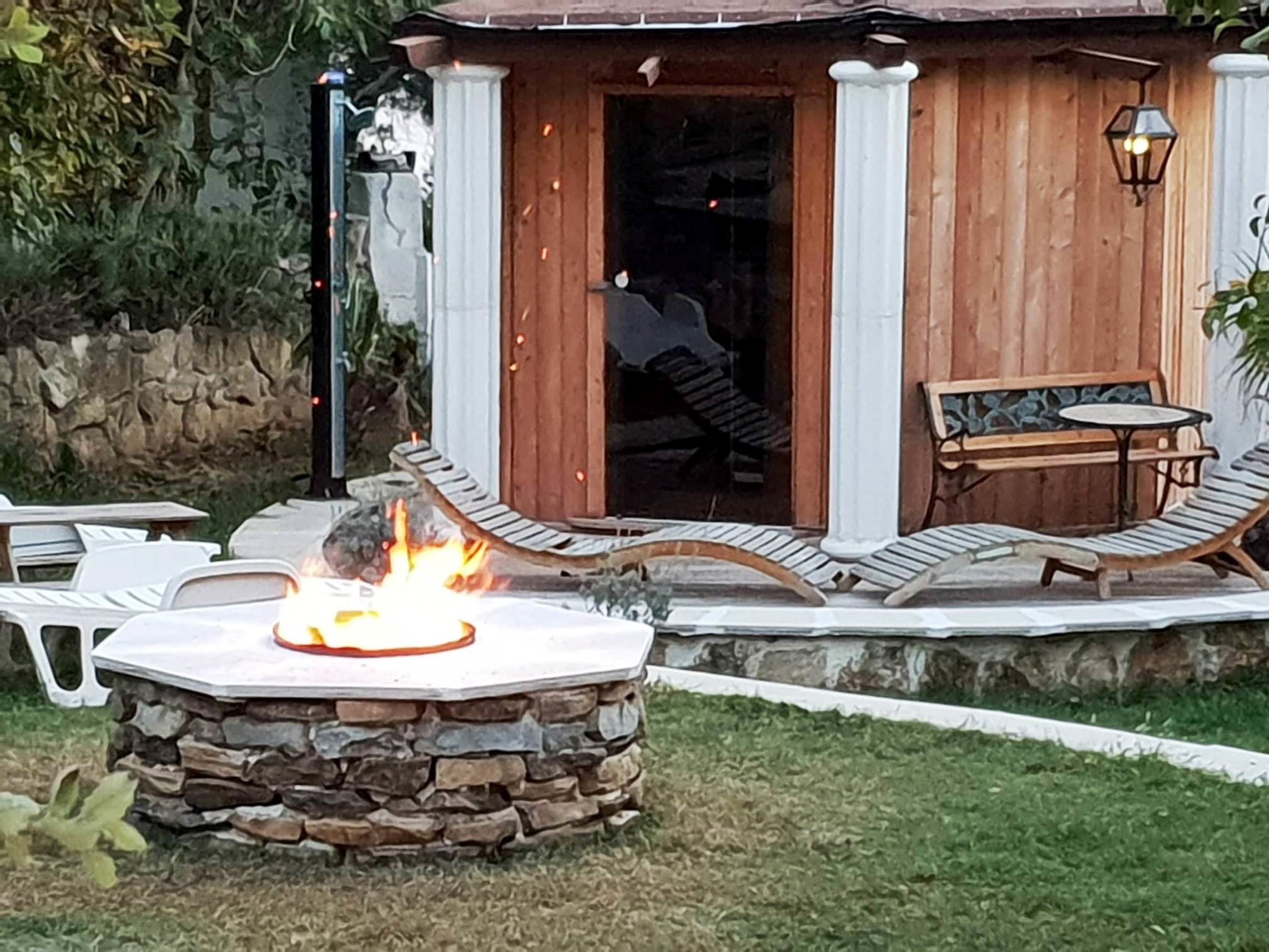 A stone fire pit with a fire, a wooden deck with a table and chairs, and a stone wall in the background.