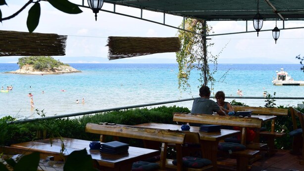 An outdoor seating area with wooden tables and benches, overlooking a beach with people swimming and a boat in the distance. - Villa Agata Rab - Apartments (Rab)