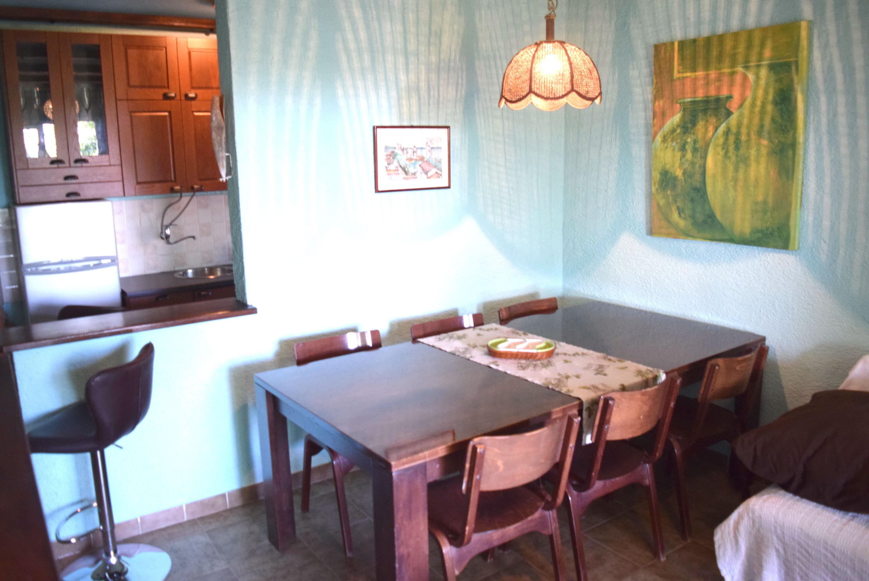 A dining area with a wooden table and chairs, a hanging light fixture, and a painting on the wall.
