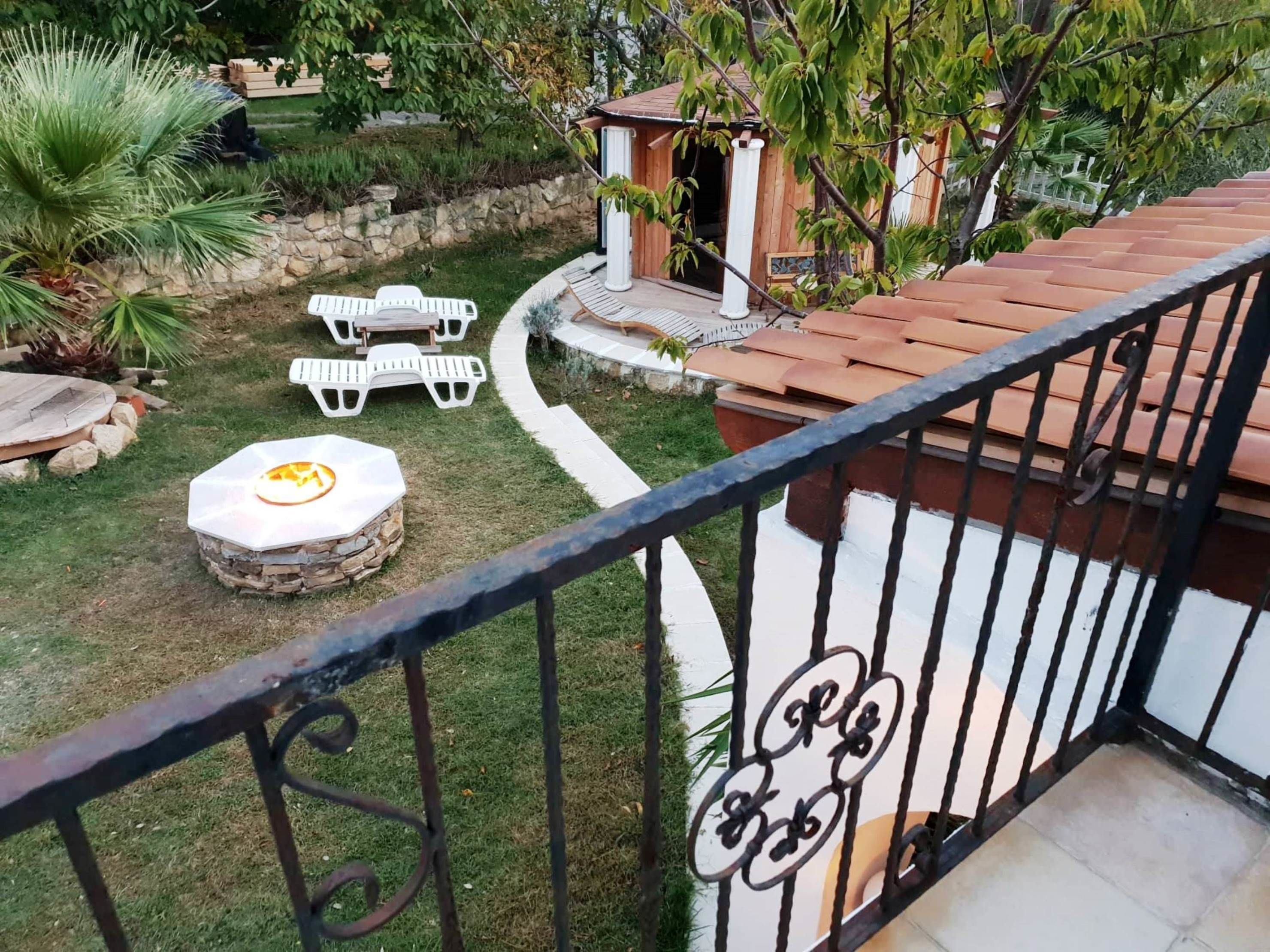 A balcony with a stone fire pit, white outdoor furniture, and a garden with various plants and a wooden structure.