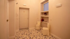 Un pasillo estrecho con una puerta cerrada, una silla blanca pequeña, un estante con una planta en maceta y un piso de baldosas.
