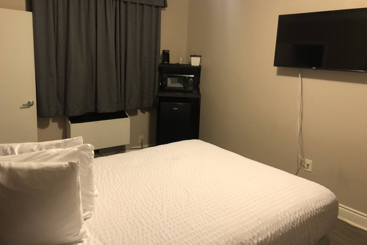 A hotel room with a bed, a television mounted on the wall, a minibar, and a window with curtains.