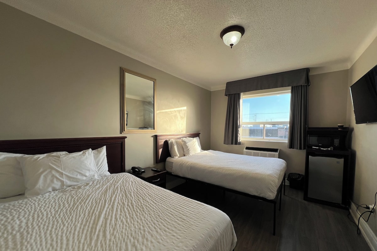 A hotel room with two beds, a desk, a mirror, a window with curtains, a TV, and a microwave.