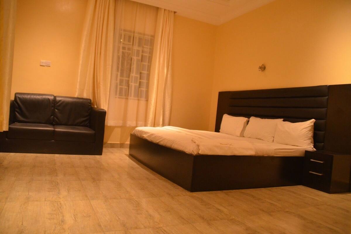 Un dormitorio con cama, sofá oscuro, ventana con cortinas y una lámpara fijada a la pared.
