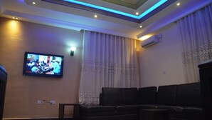 A living room with a black leather sofa, a flat-screen TV mounted on the wall, and a ceiling with blue LED lighting. - House X (Abuja)