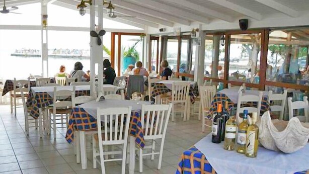 An outdoor restaurant with white tables and chairs, covered by a roof, and bottles of wine on the tables. - Corali Studios (Agios Vasileios)