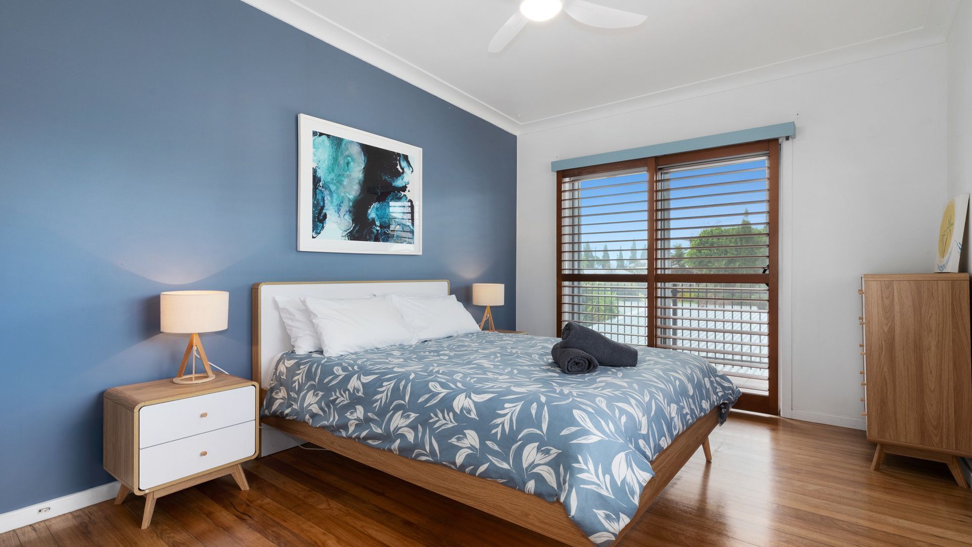 A bedroom with a bed, bedside tables, a ceiling fan, a window with blinds, and a wooden cabinet.