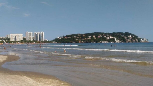 Une plage où l’on voit des gens se baigner et se promener le long du rivage, des bâtiments en arrière-plan et une colline parsemée de maisons.