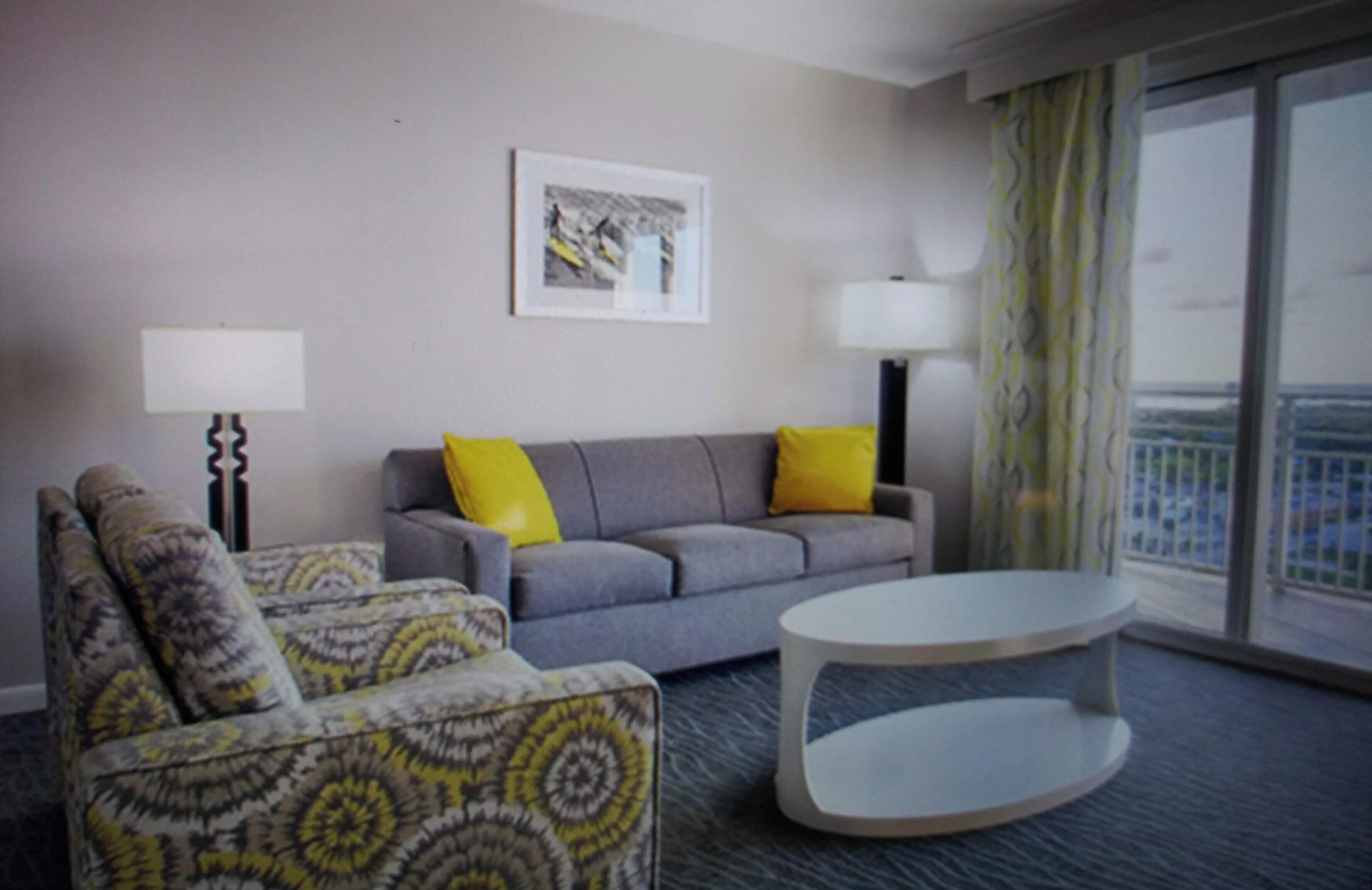 A bedroom living room with a grey sofa, patterned armchair, and a white coffee table. There are two lamps, a framed picture on the wall, and a view of the city through large windows.