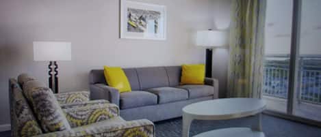 A bedroom living room with a grey sofa, patterned armchair, and a white coffee table. There are two lamps, a framed picture on the wall, and a view of the city through large windows.