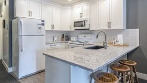Cocina moderna con armarios blancos, microondas empotrado, refrigerador, pileta y encimera de granito. Hay tres taburetes de madera en la isla.