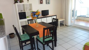 En matplats med ett bord, stolar, en vas med blommor, en tv och ett vitt skåp med hyllor.