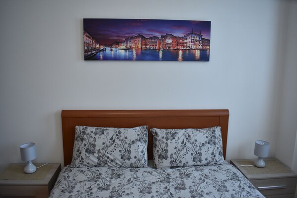 A hotel room with a bed, floral-patterned pillows, a nightstand with a lamp, and a painting of a cityscape on the wall. - Elegant and Large apartment with free car parking near Venice (Venezia)