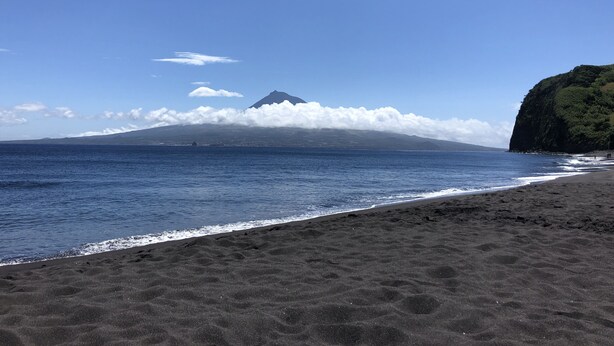 A beach with dark sand, a clear blue sky, and a mountain with clouds in the background. - Kings' Retreat   (Horta)