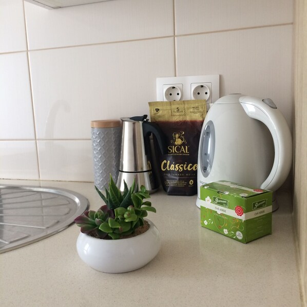 Un taulell de cuina amb una cafetera, un paquet de cafè, una caixa de te, una bullidora i una petita planta en test.
