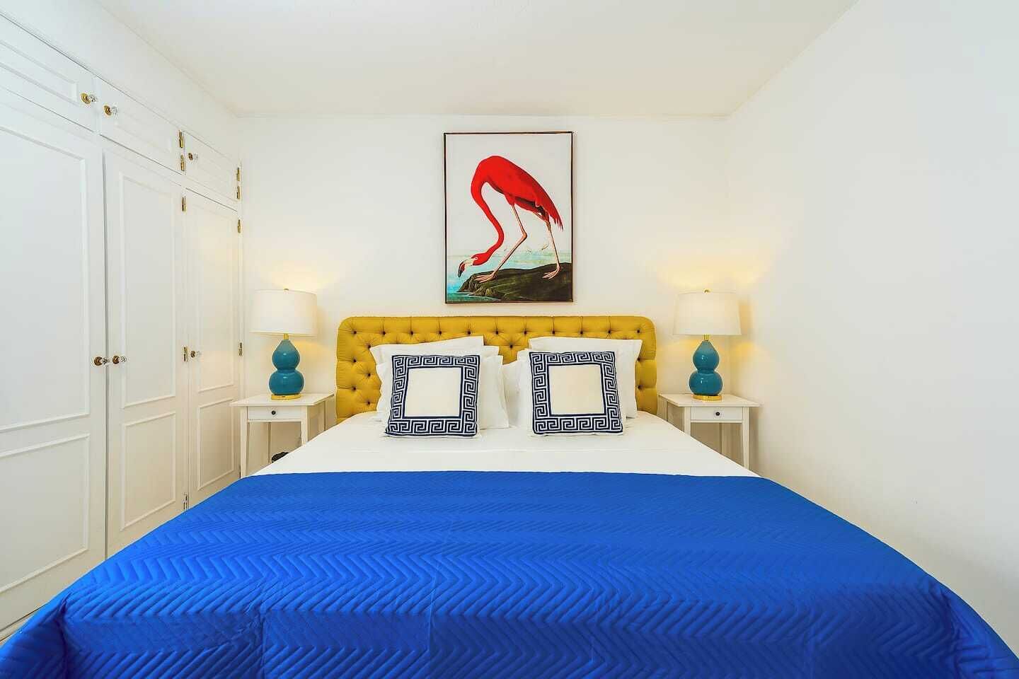 Ein Schlafzimmer mit einem gelben, gesteppten Kopfteil, einer blauen Bettdecke und zwei passenden Kissen. An der Wand hängt ein gerahmtes Bild eines Flamingos.