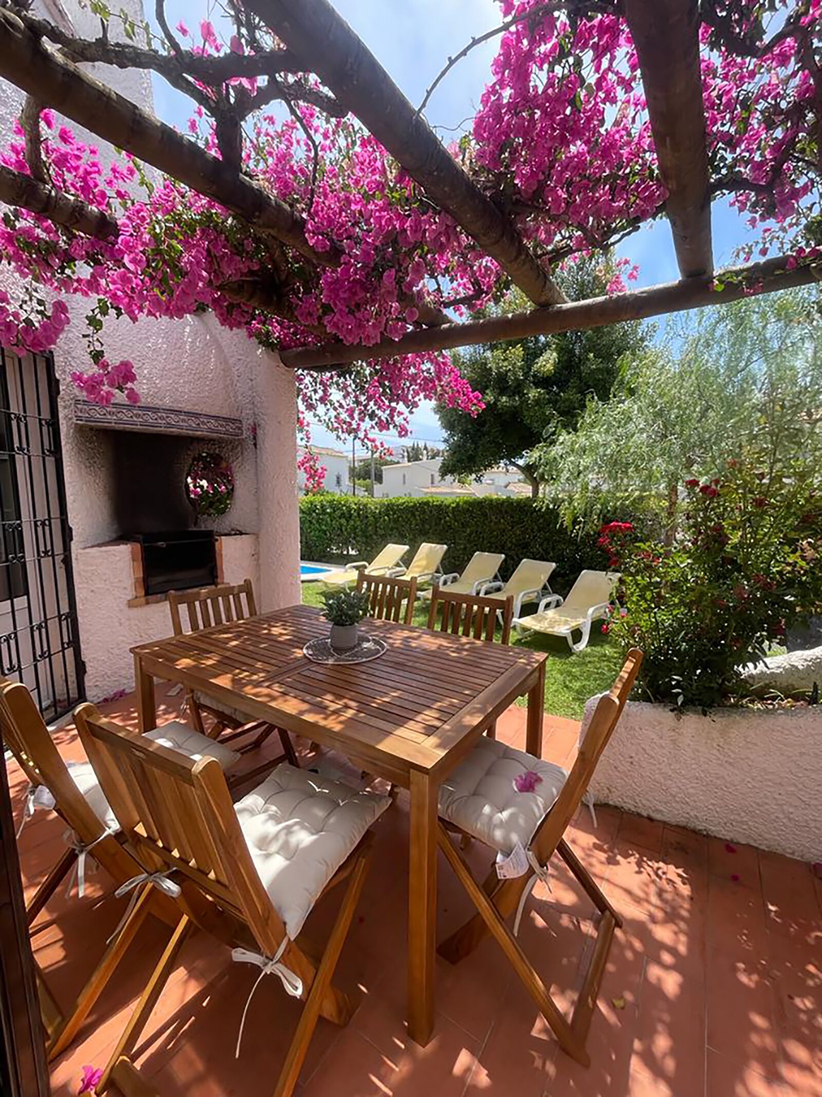 A patio with a wooden table and chairs, surrounded by pink flowering trees.