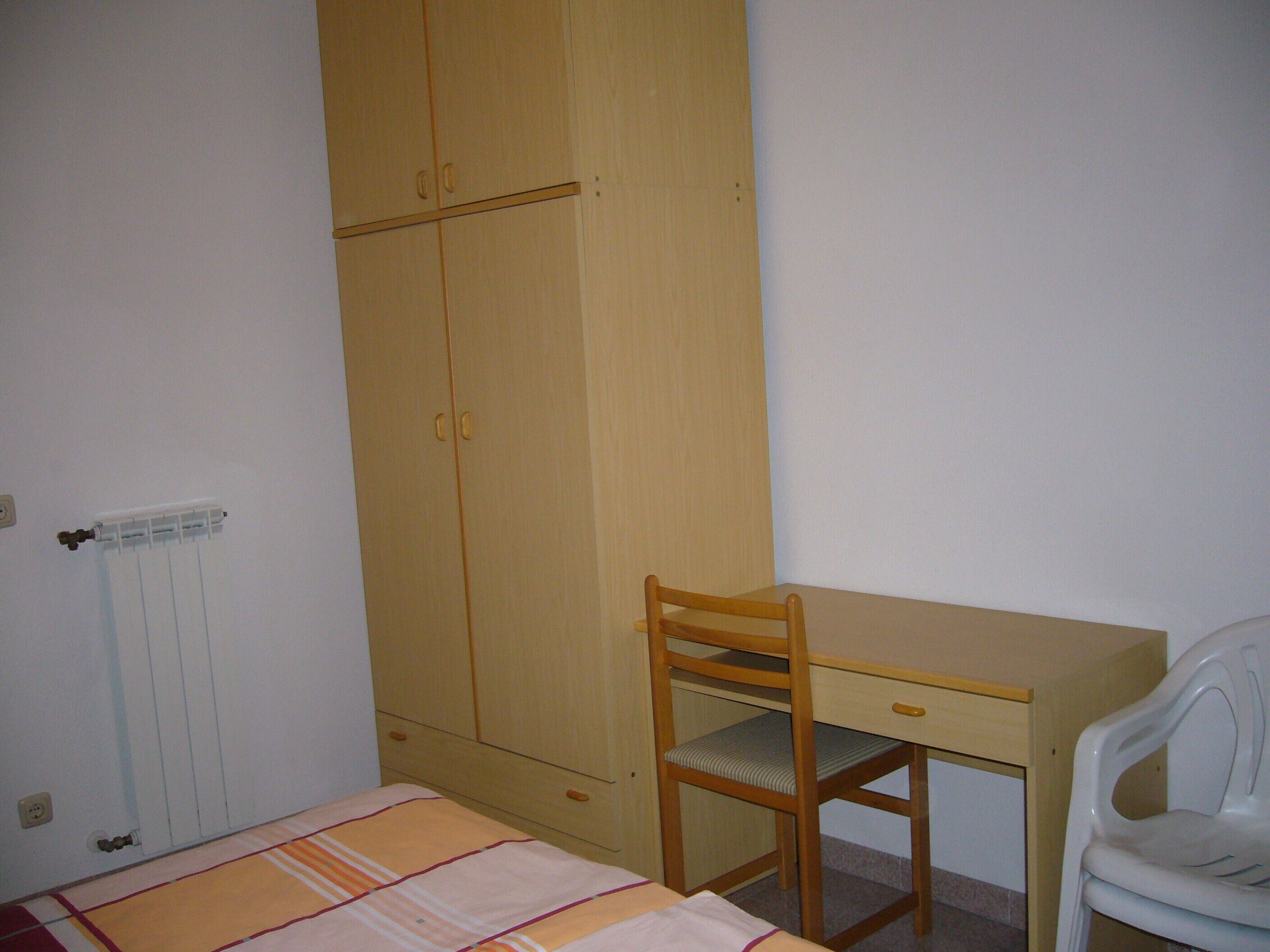A bedroom with a wooden wardrobe, a desk with a chair, and a bed with a striped bedspread.