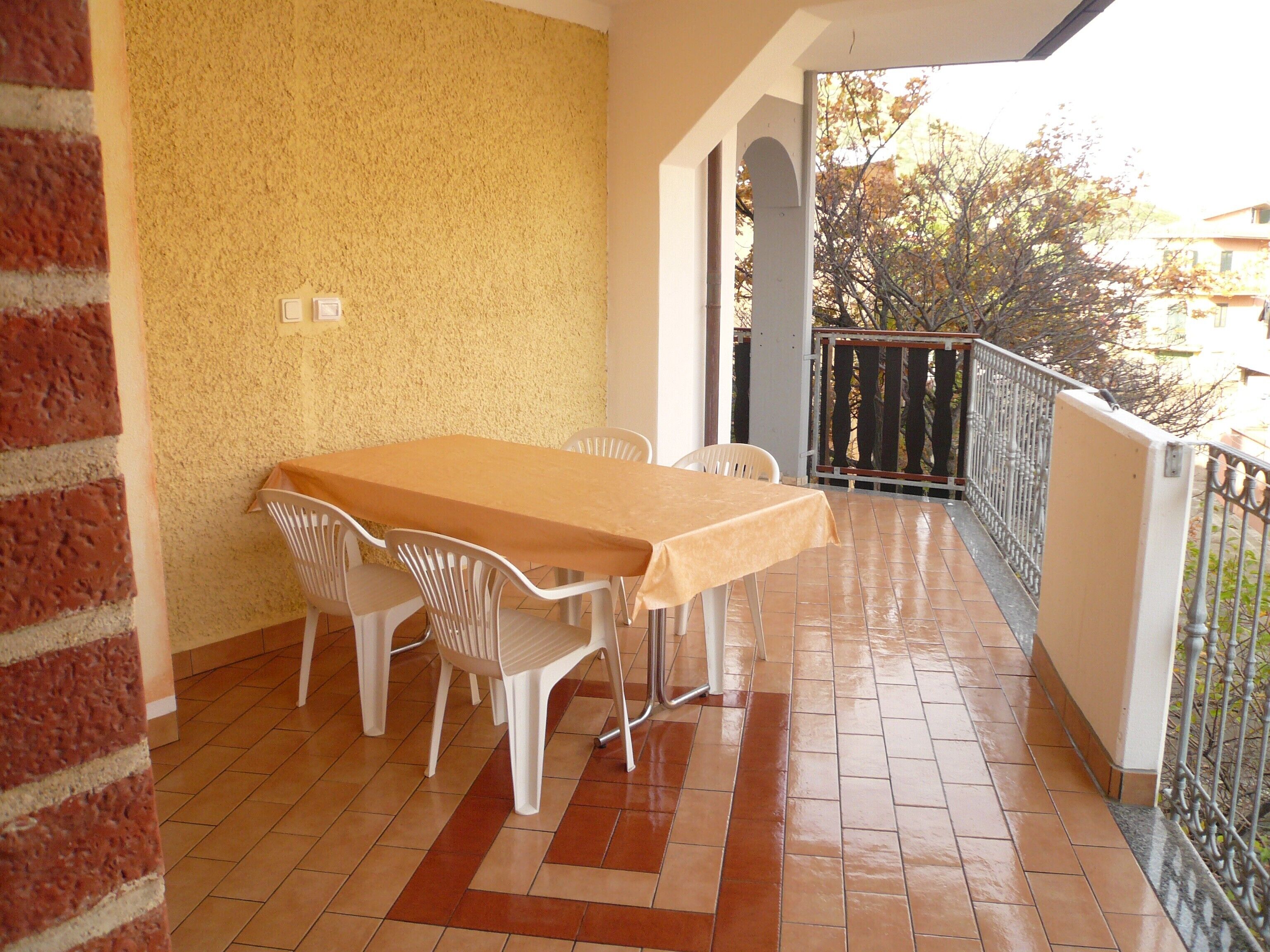 A covered patio with a tiled floor, a table with chairs, and a view of trees and buildings in the background.
