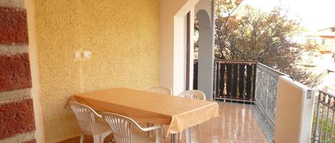 A covered patio with a tiled floor, a table with chairs, and a view of trees and buildings in the background.