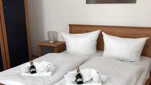 Una cama doble con ropa blanca, dos almohadas y dos botellas de champán sobre una toalla. En la pared, un cuadro de una puesta de sol con pilares de piedra.