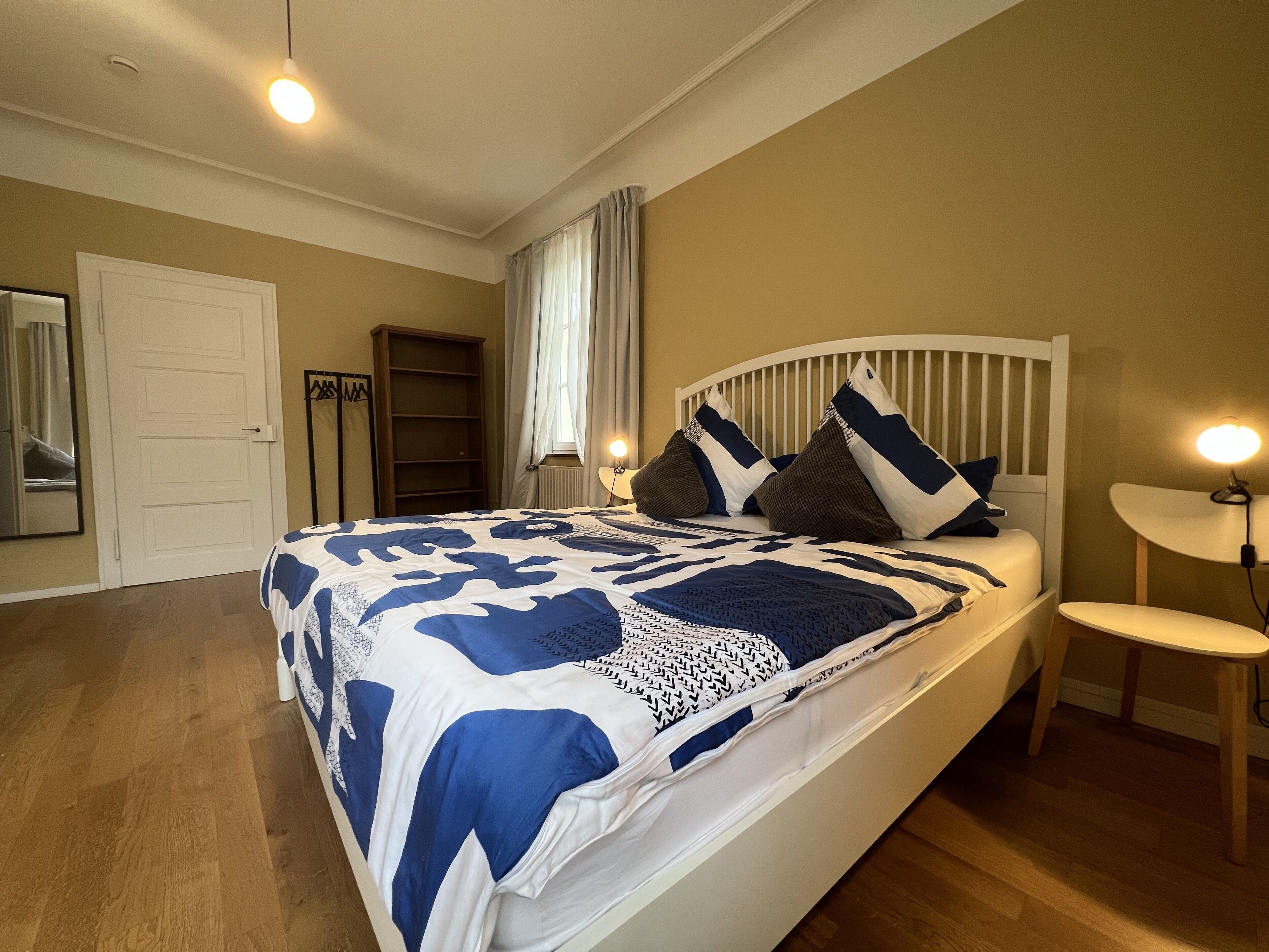 A bedroom with a bed, bedside tables, a nightstand, a mirror, a wardrobe, and a window with curtains.