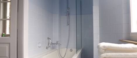 Un baño con bañera, ducha y zona de ducha con cerramiento de vidrio. Hay azulejos blancos en las paredes y una ventana que proporciona luz natural.