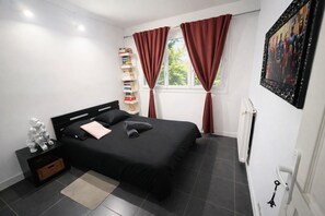 A bedroom with a black bed, a bedside table with a lamp, a bookshelf, and a window with red curtains. - Appt Cosy Porte Paris Bercy / Parc Floral de Vincennes + Parking (Saint maurice)