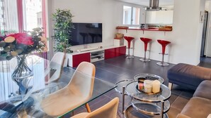 Un soggiorno moderno con tavolo da pranzo in vetro, comodi posti a sedere, angolo cottura con sgabelli da bar rossi e un grande televisore.