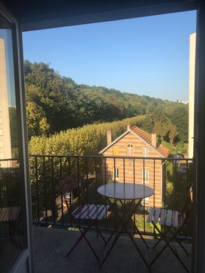 A balcony with a table and chairs, overlooking a brick building and a forested hillside. - Appt Cosy Porte Paris Bercy / Parc Floral de Vincennes + Parking (Saint maurice)
