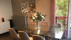 Un'area pranzo moderna con tavolo in vetro, quattro sedie, un vaso di fiori, un termosifone e un elemento decorativo a parete.