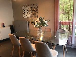 A modern dining area with a glass table, four chairs, a vase of flowers, a radiator, and a decorative wall piece. - Appt Cosy Porte Paris Bercy / Parc Floral de Vincennes + Parking (Saint maurice)