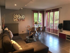 A modern living room with a sofa, a dining table with chairs, a TV, and large windows with curtains. - Appt Cosy Porte Paris Bercy / Parc Floral de Vincennes + Parking (Saint maurice)