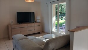 A modern living room with a sofa, a TV on a wooden cabinet, a small table, and a view of an outdoor garden.