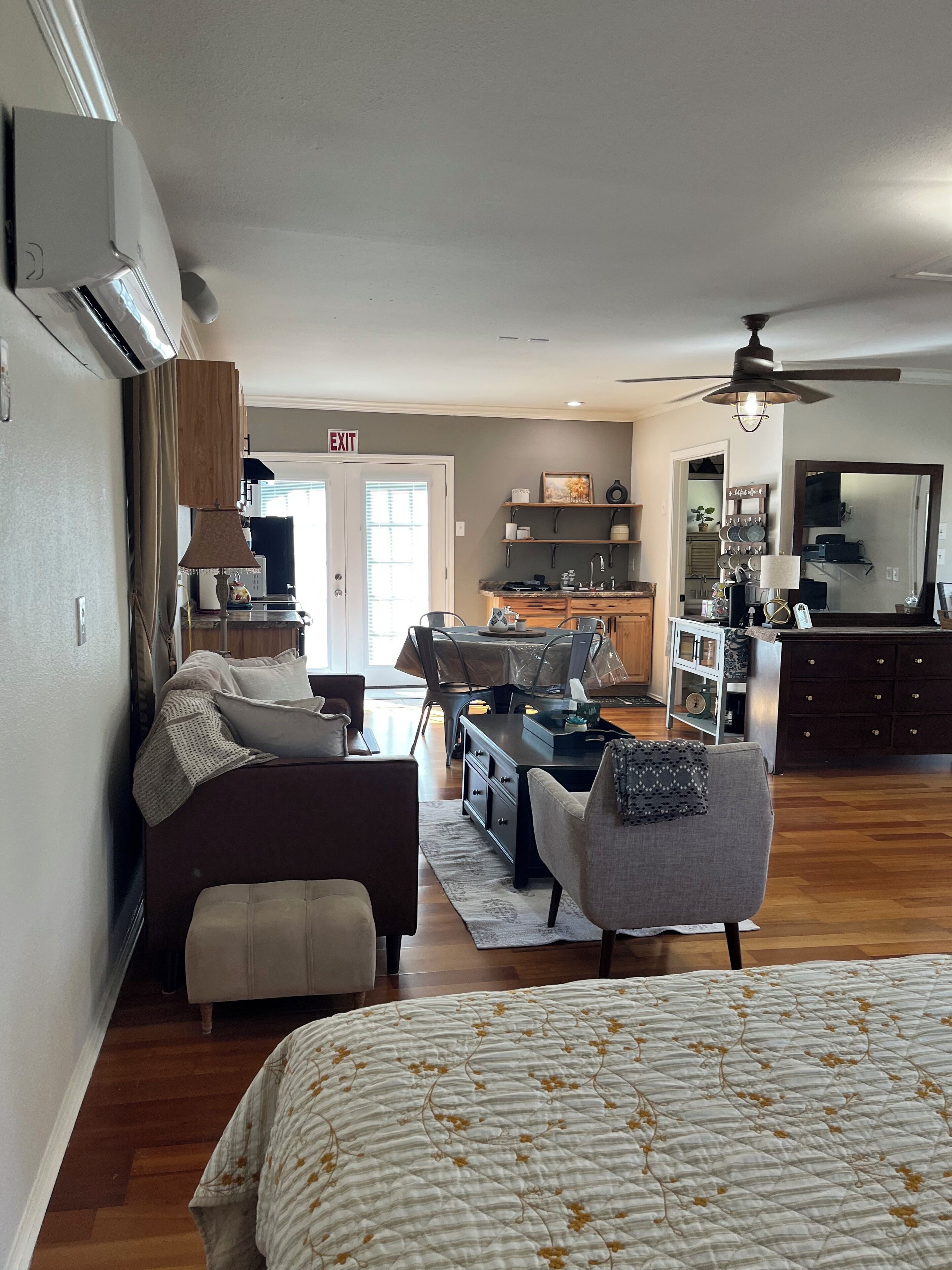 A spacious living area with a sofa, armchair, and a bed. There is a kitchen area with a dining table and chairs. An exit sign is visible on the door.