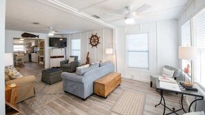 Un vaste espace de vie comprenant un canapé, un pouf et une table basse en bois. Un téléviseur est fixé au mur, il y a également un ventilateur de plafond et une fenêtre équipée de stores.