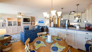 A modern kitchen with white cabinets, a dining table with a floral table runner, and a blue sofa in the living area. - Sur La Plage-Oceanfront & Pool (Fernandina Beach)
